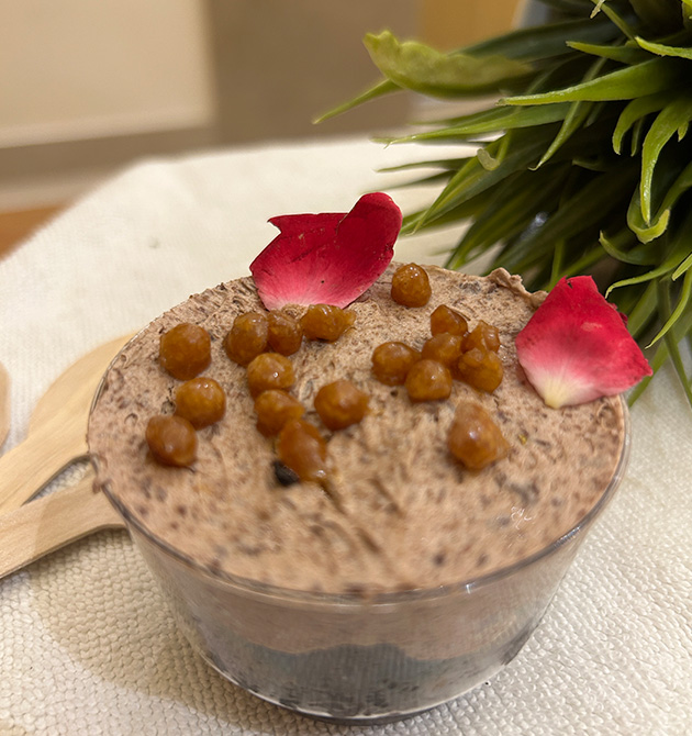 A dark whipped dessert in a glass bowl topped with crisp golden boondi and rose petals.