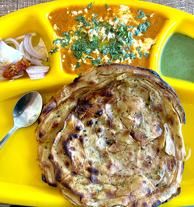 A lachha paratha with paneer butter masala, green chutney and sliced onion on a yellow tray.
