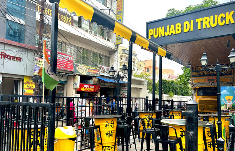 The open-air Punjab Di Truck restaurant at street level with yellow drum tables under a striped canopy.