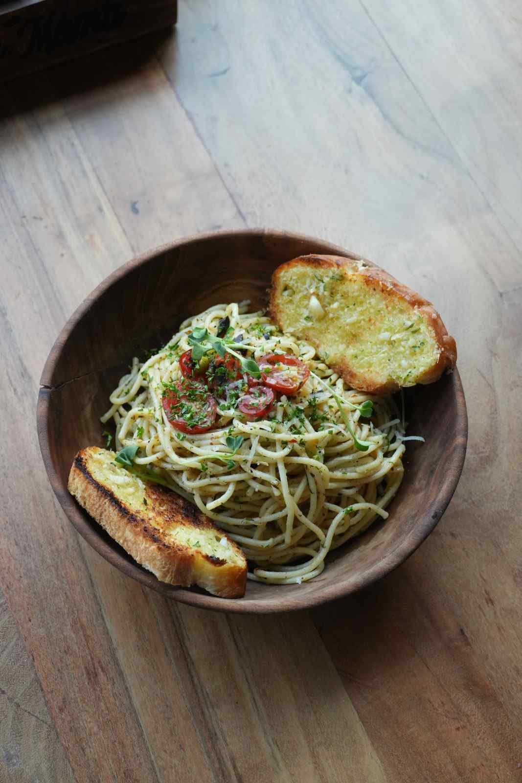 A bowl of green pesto spaghetti with garlic bread on a wooden table.