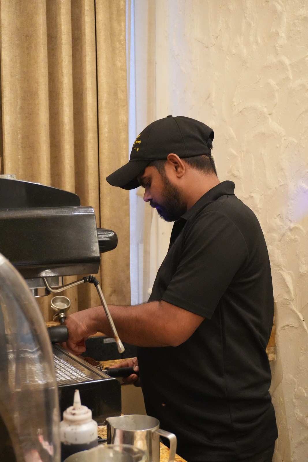 A barista in a black cap steaming milk at the espresso machine.
