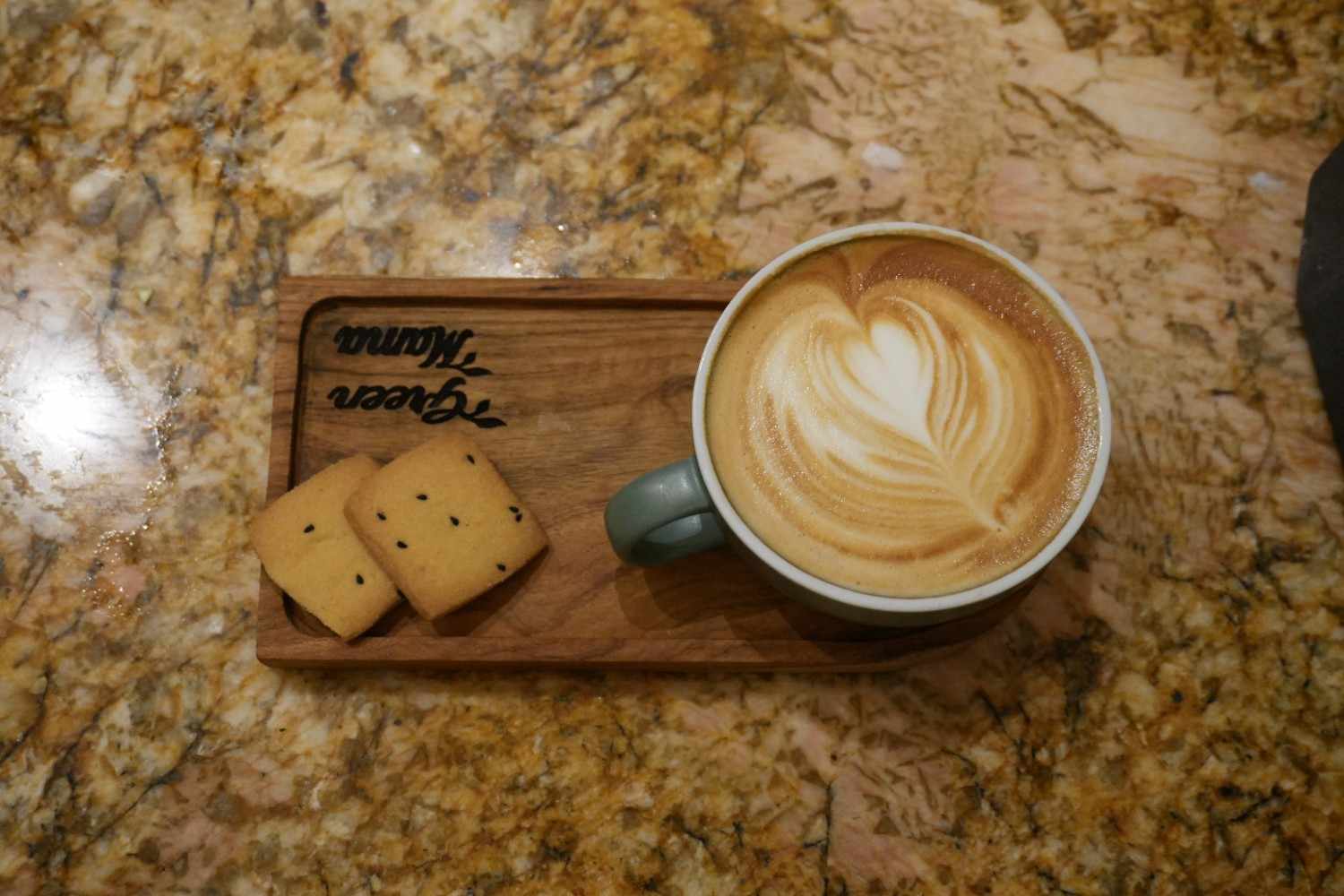 A latte with rosetta foam art on a wooden board with two biscuits.