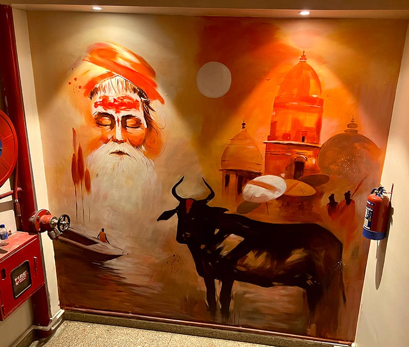 A wall mural in orange tones of a sadhu, a black bull and temple domes above a boat.
