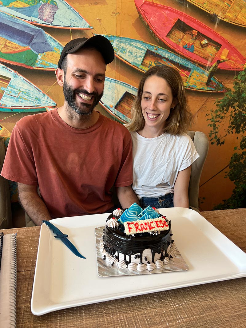 A couple smiling at a chocolate birthday cake iced with a name, in front of the painted wall of boats.
