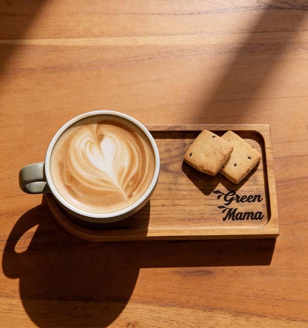 A latte with heart-shaped foam art and two biscuits on a wooden Green Mama board.
