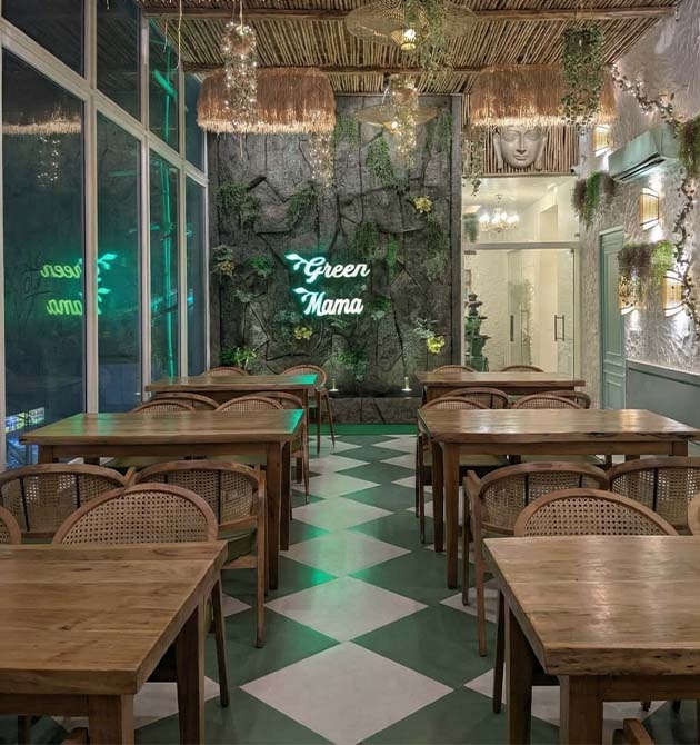Tables at Green Mama in front of a planted stone wall lit by a green neon sign.