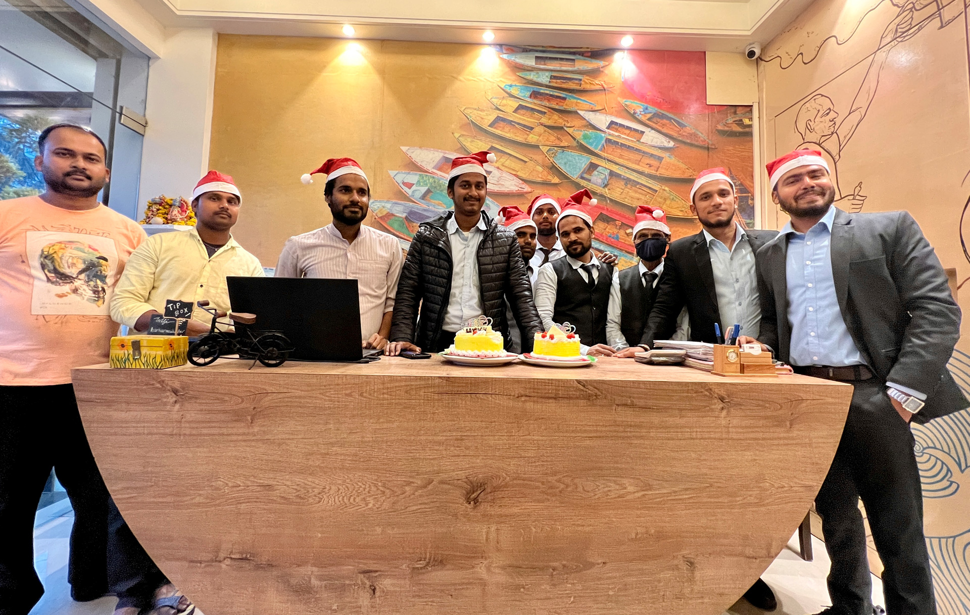 The hotel team behind the reception desk in Santa hats, with two cakes, in front of the painted wall of boats.